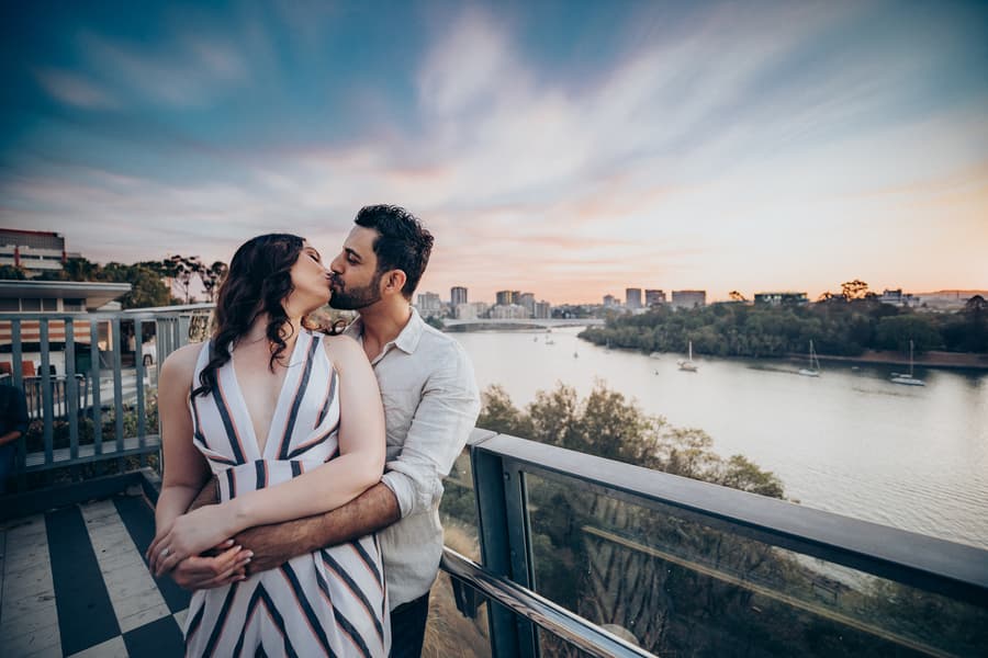 The couple embraces and kisses with a scenic river and city skyline in the background at Kangaroo Point, Brisbane.