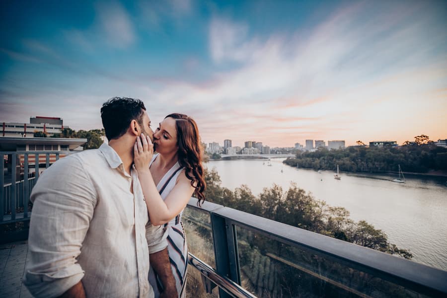 The couple shares a tender kiss with a river and cityscape in the background at Kangaroo Point, Brisbane.