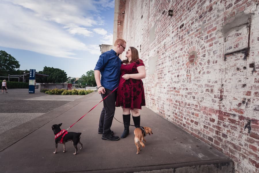 The couple shares a tender moment, standing close with their dogs, against a brick wall.