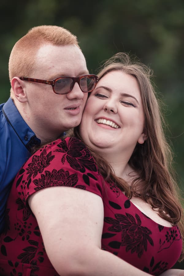The couple embraces with smiles, surrounded by greenery, creating a warm and intimate atmosphere.