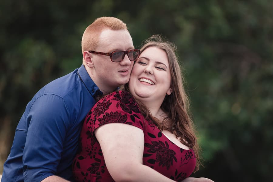 The couple embraces with smiles, surrounded by greenery, creating a warm and intimate atmosphere.