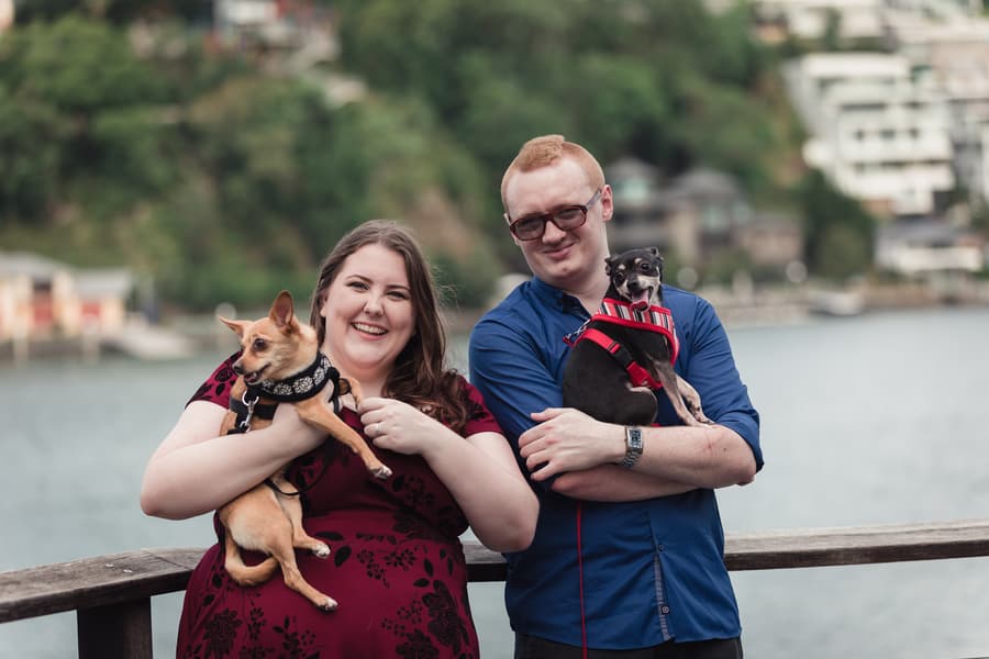 The couple stands by a river, each holding a dog, with a scenic view of trees and water behind them.