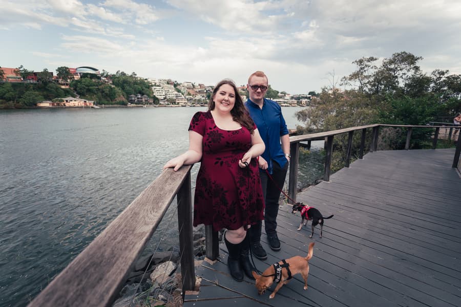The couple leans on a wooden railing by the river, with their dogs exploring nearby.