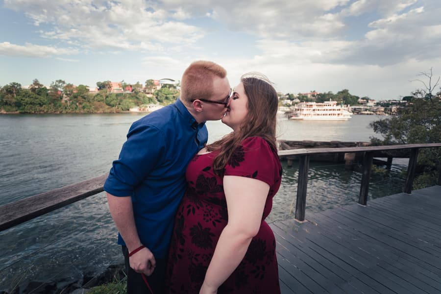The couple shares a kiss on a wooden walkway by the river, under a partly cloudy sky.