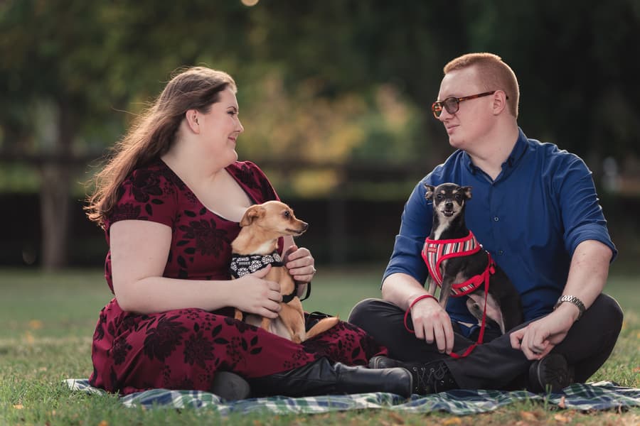 The couple sits on a blanket in a grassy area, each holding a dog, looking at each other warmly.