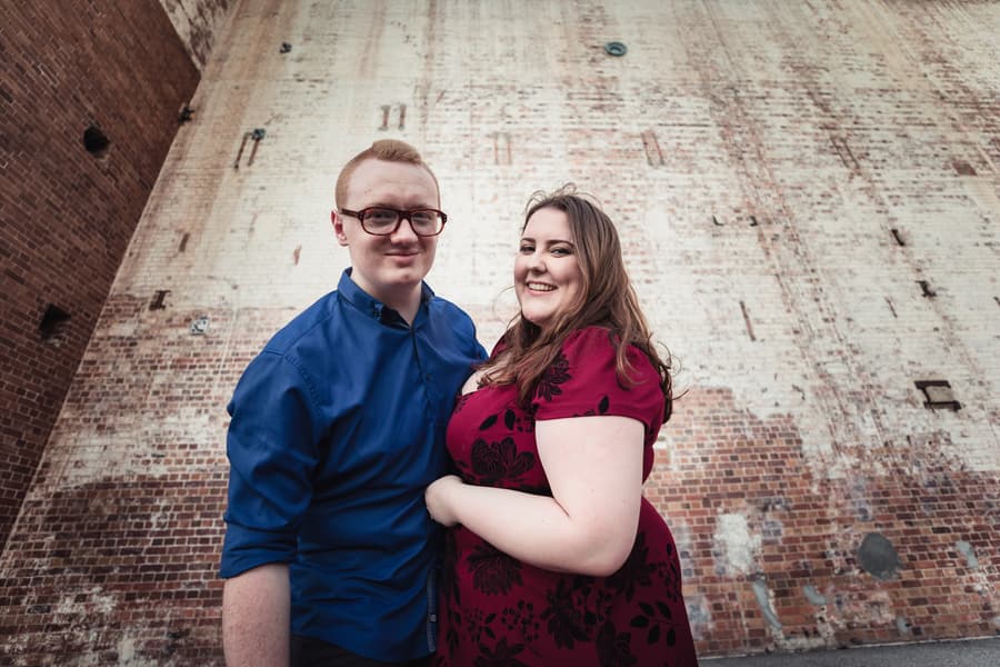 The couple stands close together against a large brick wall at Brisbane Powerhouse, smiling warmly.
