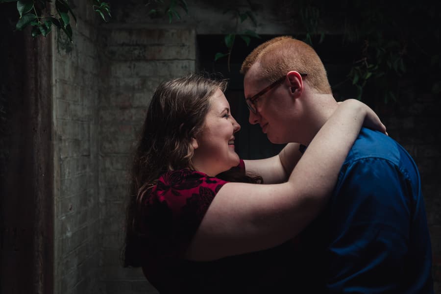 The couple embraces closely in a dimly lit corner at Brisbane Powerhouse, smiling softly at each other.