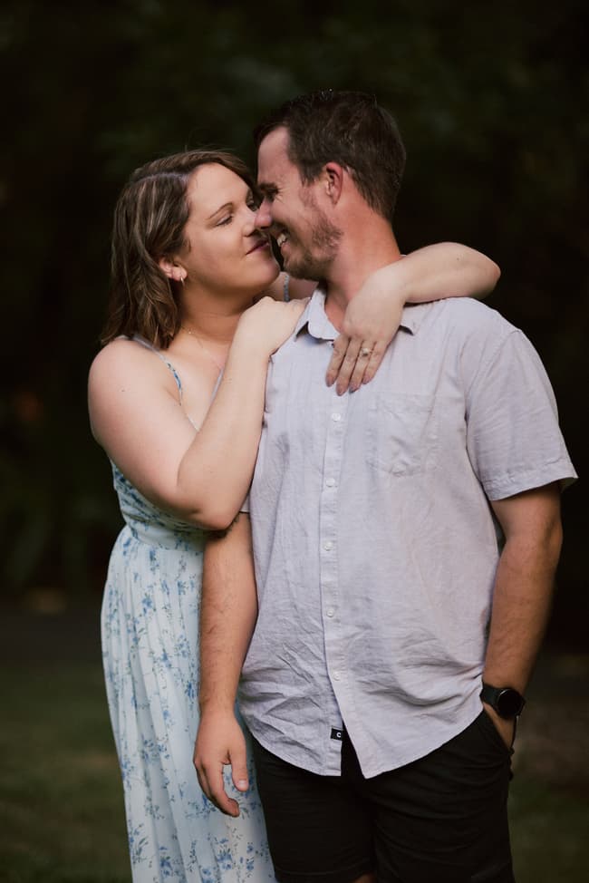 The couple embraces closely, with one leaning on the other's shoulder, in a shaded area.