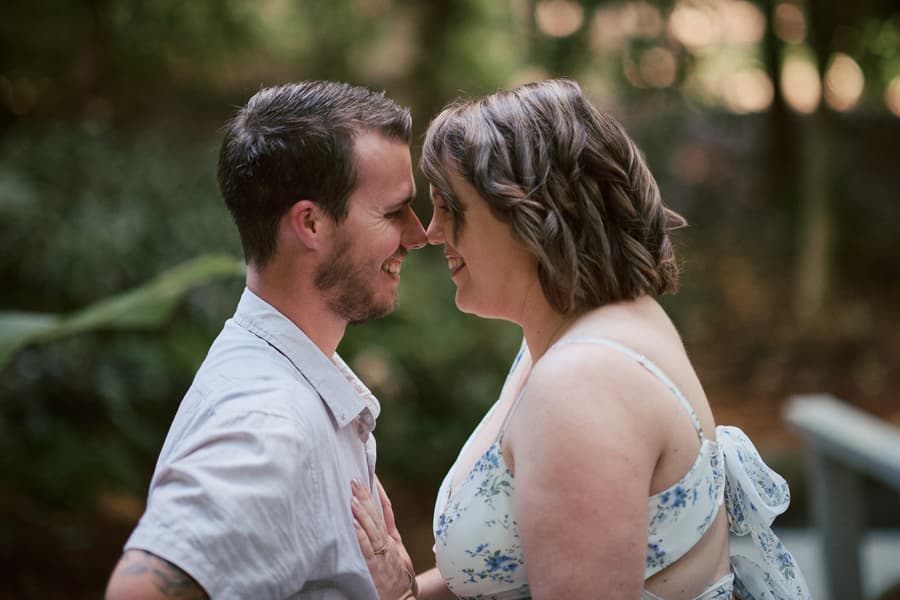 The couple stands close, smiling at each other, with a forest backdrop.