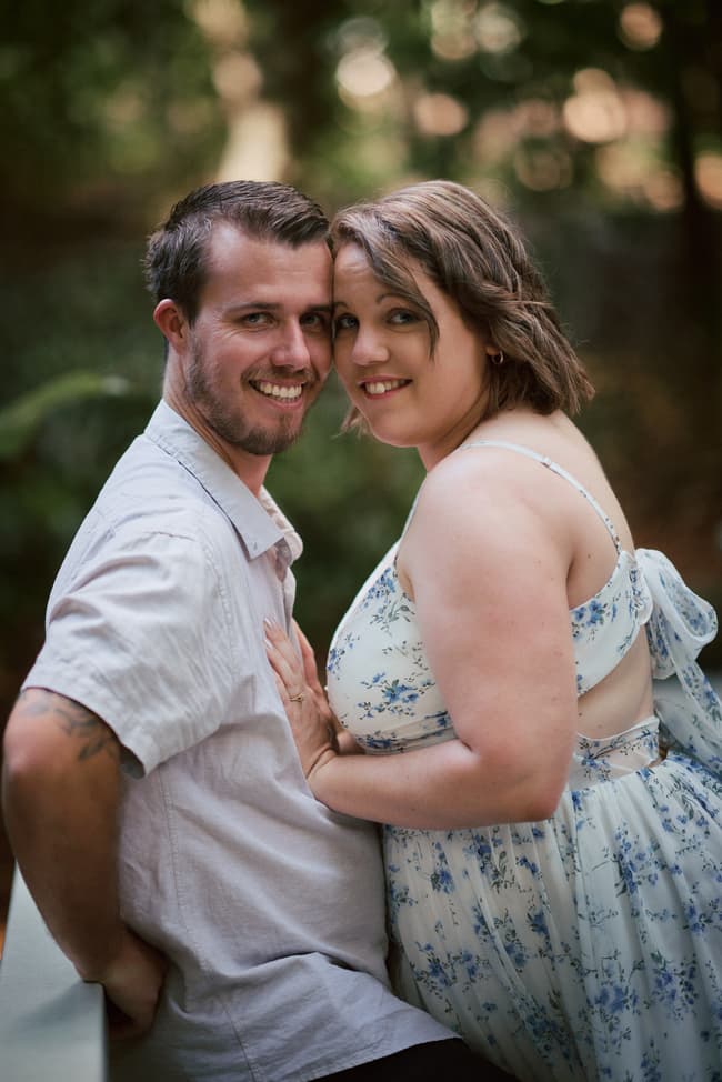 The couple leans against a bridge railing, smiling warmly at the camera.