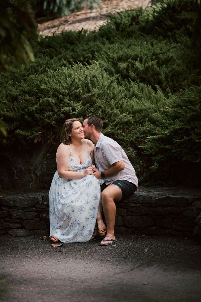 The couple sits on a stone bench, one kissing the other's cheek, surrounded by greenery.