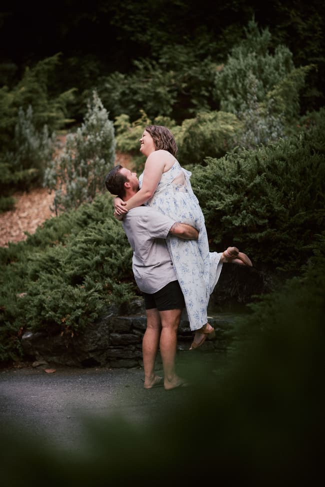 One person lifts the other playfully in a garden setting, both smiling, surrounded by dense foliage.
