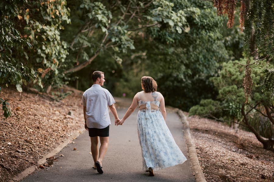 The couple walking hand in hand down a tree-lined path, with leaves scattered on the ground at Brisbane Botanic Gardens.