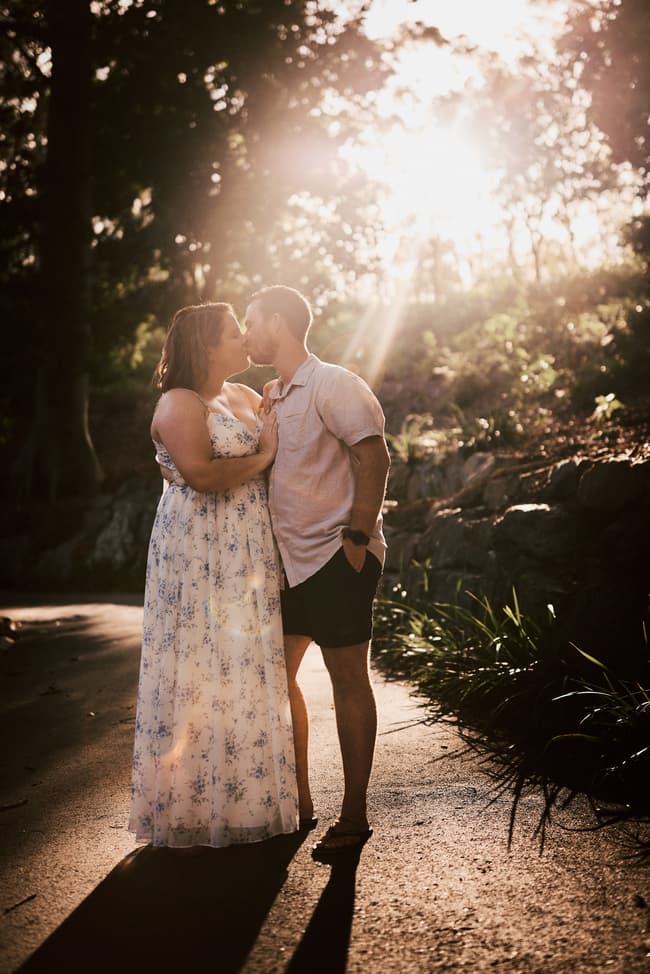 The couple shares a kiss on a sunlit path, with rays of light filtering through the trees.
