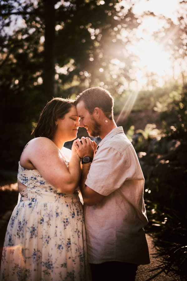 The couple stands close, foreheads touching, bathed in warm sunlight with trees in the background.