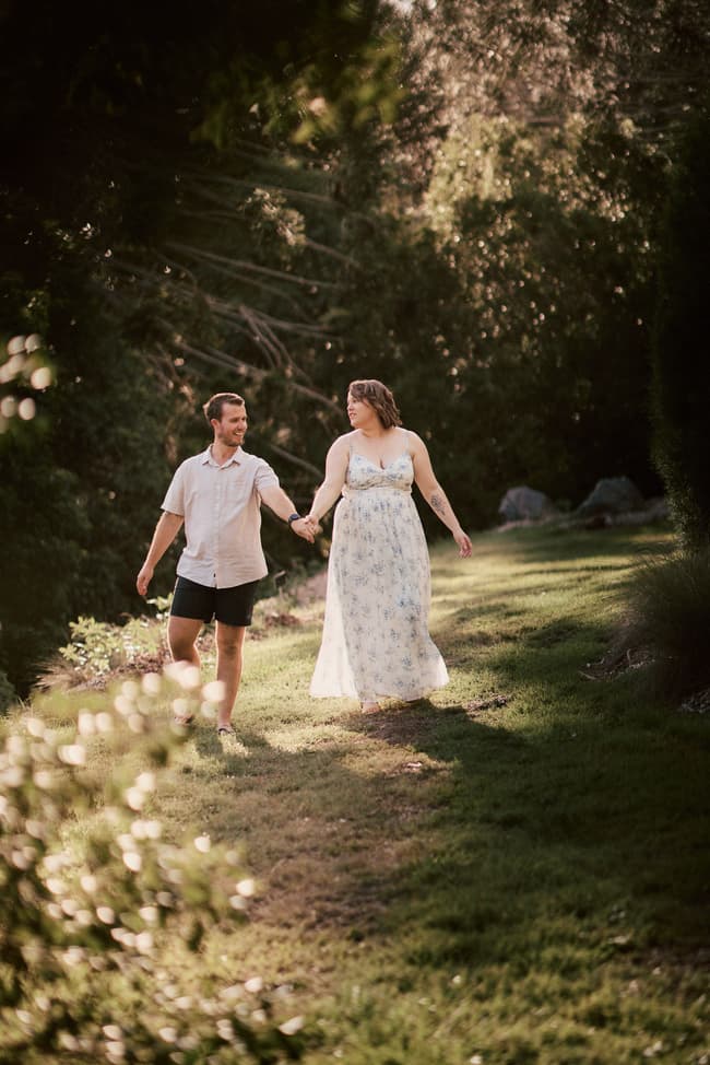 The couple walking hand in hand on a grassy path, surrounded by trees and soft sunlight.