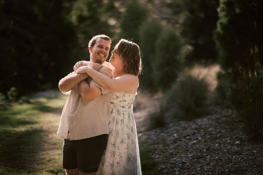 One person embraces the other from behind, both smiling, with a backdrop of trees and soft light.