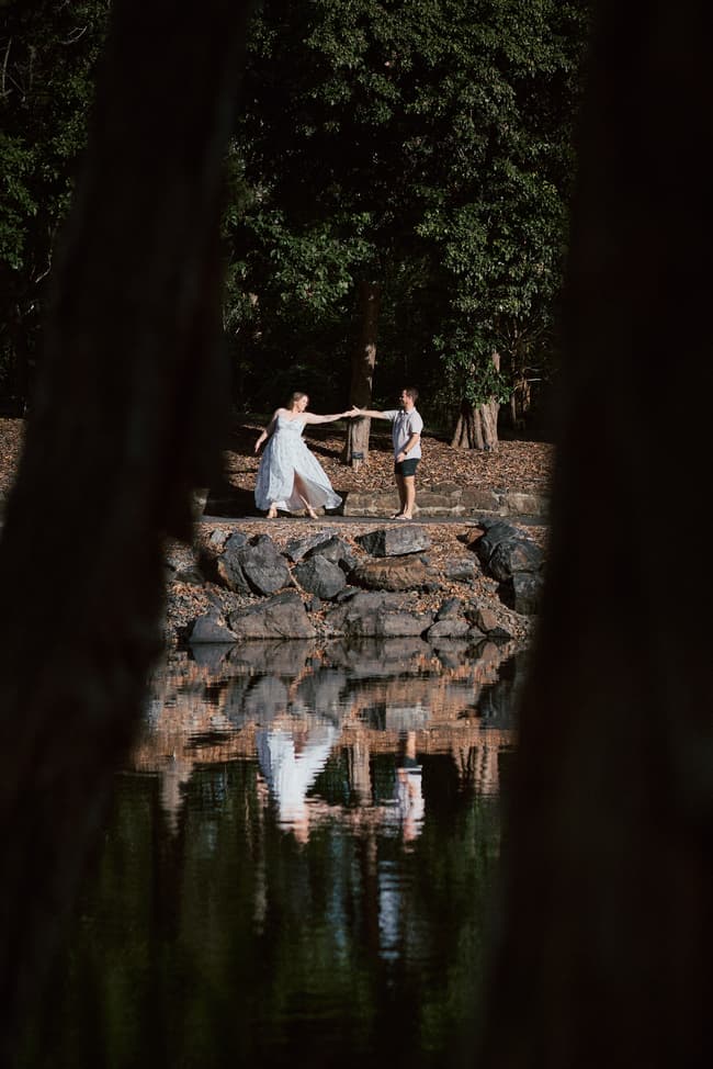 The couple stands on rocks by a pond, holding hands, with trees reflected in the water.