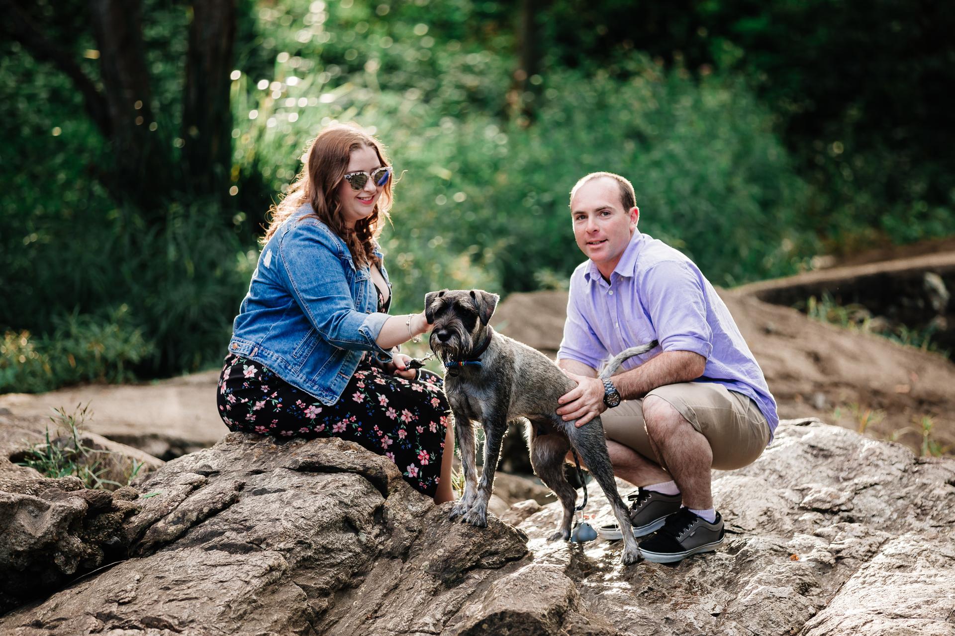 The couple kneels on rocks with their dog at Merv Ewart Reserve, surrounded by greenery.