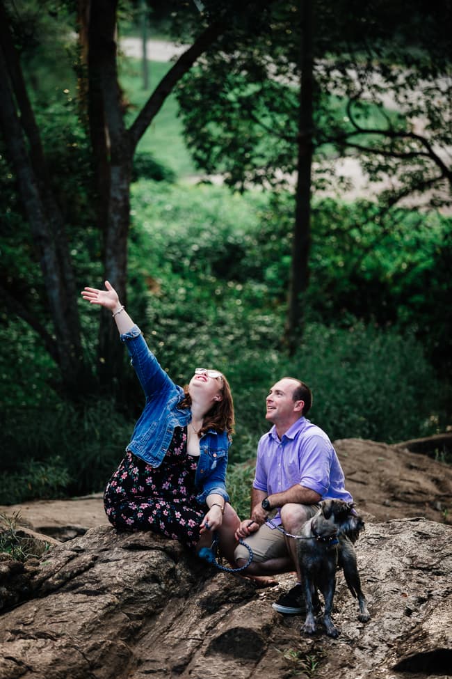 The couple sits on rocks, looking up joyfully, with trees in the background at Merv Ewart Reserve.