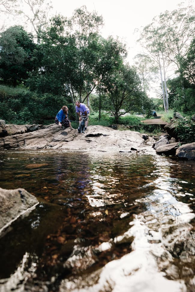The couple is by a stream, surrounded by trees, with reflections in the water at Merv Ewart Reserve.