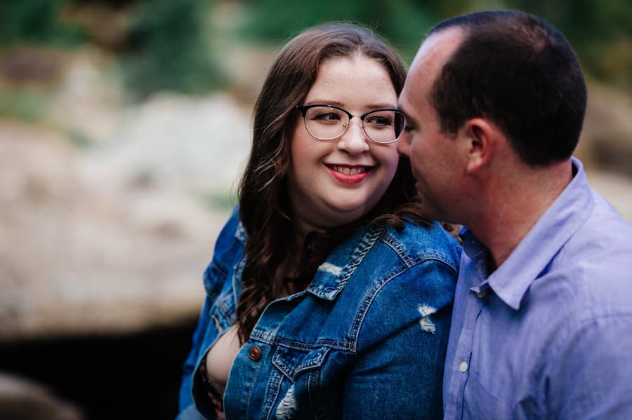 The couple shares a warm glance, sitting on rocks with greenery in the background at Merv Ewart Reserve.