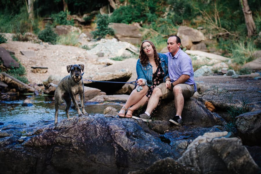 The couple sits on rocks by a stream, with their dog on a leash, surrounded by nature at Merv Ewart Reserve.