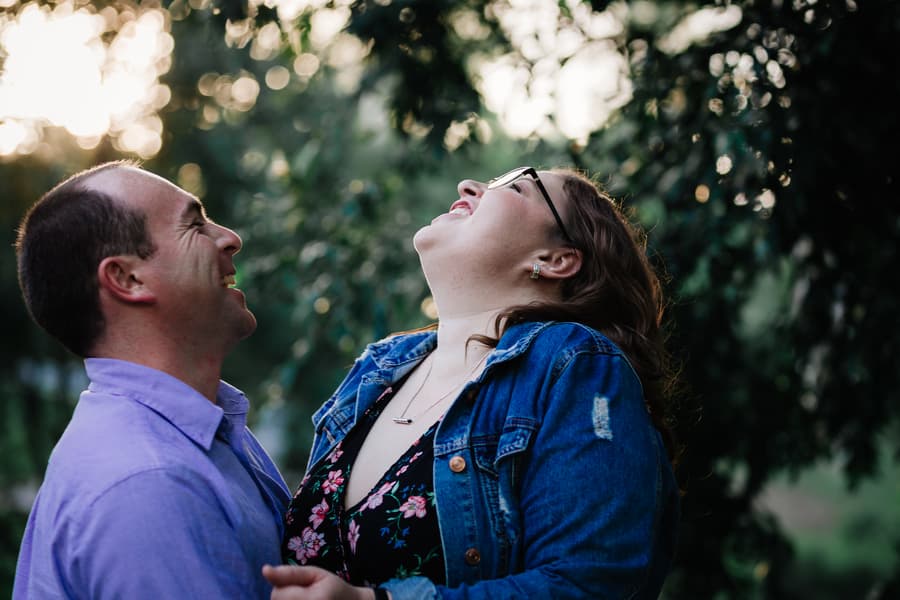 The couple laughs together under the trees, with sunlight filtering through at Merv Ewart Reserve.