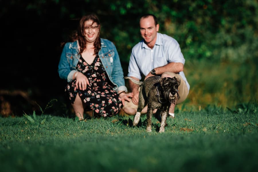 The couple kneels on grass, smiling as their dog runs towards the camera at Merv Ewart Reserve.