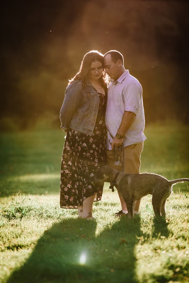 The couple stands closely with a dog at Merv Ewart Reserve, bathed in warm sunlight.