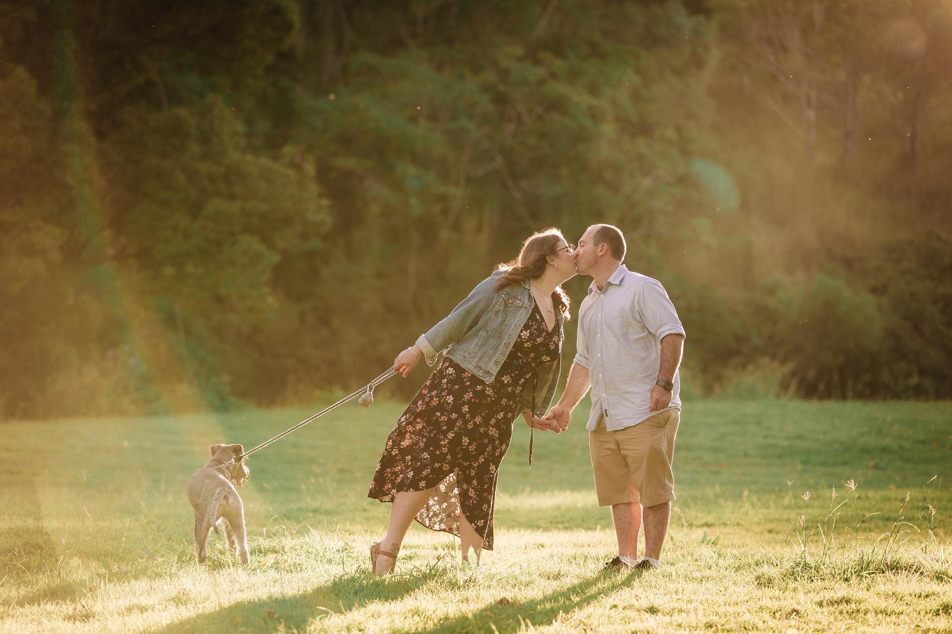 The couple kisses while holding hands and a dog leash in a sunlit park setting.