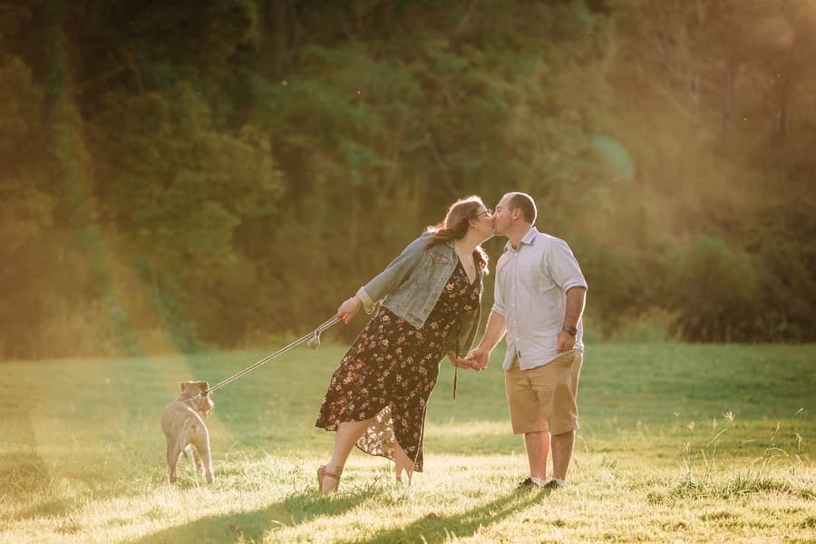 The couple kisses while holding hands and a dog leash in a sunlit park setting.