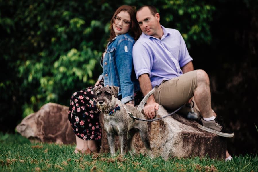 The couple sits on a rock with their dog, surrounded by lush greenery.