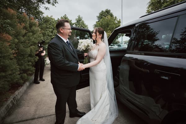 The bride Terion, holding a bouquet, stands outside a black vehicle holding hands with an older man, possibly her father, at Sandstone Point Hotel — The Pavilion. A photographer is visible in the background near greenery.