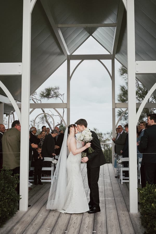 Terion and Tyler kiss at the altar during their wedding ceremony at Sandstone Point Hotel — The Pavilion, surrounded by seated guests applauding.