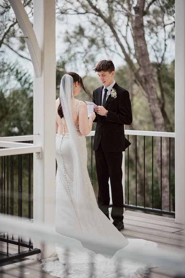 Terion the bride and Tyler the groom stand together on a balcony at Sandstone Point Hotelallo, with Terion in a white wedding gown and veil and Tyler in a black suit reading from a piece of paper.