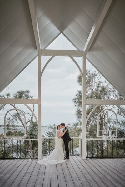 The bride Terion and groom Tyler share a kiss under a white wooden pavilion with a view of trees and water. Winter wedding light photography Brisbane by She Said Yes.