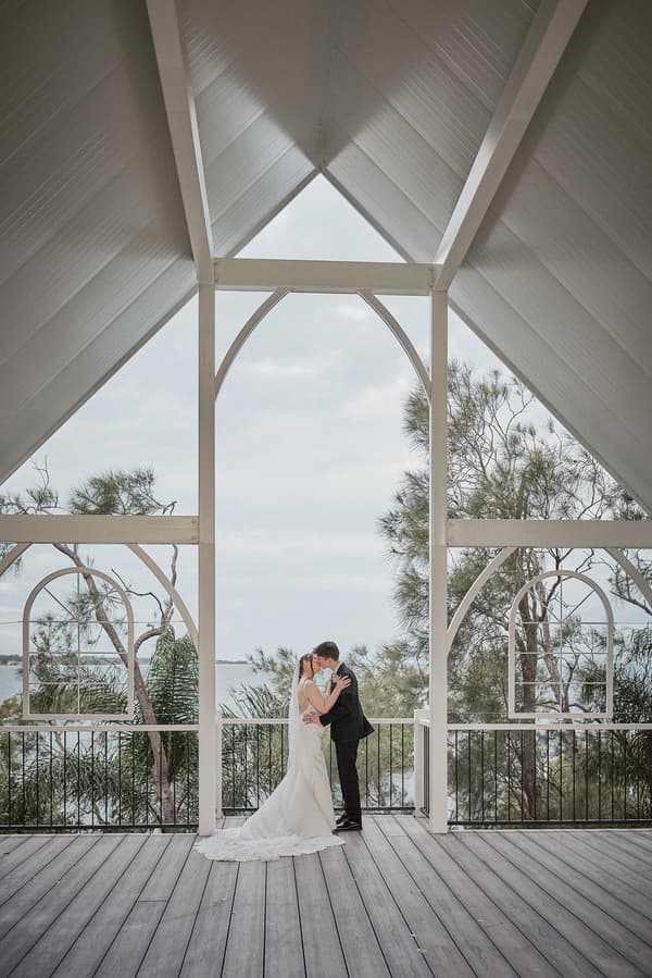 The bride Terion and groom Tyler share a kiss under a white wooden pavilion with a view of trees and water at Sandstone Point Hotel.