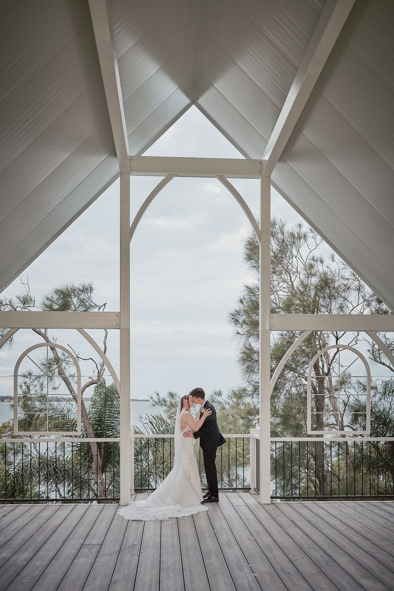 The bride Terion and groom Tyler share a kiss under a white wooden pavilion with a view of trees and water. Destination wedding photographer by She Said Yes.