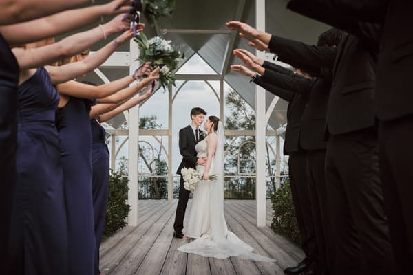 Terion and Tyler stand facing each other, framed by bridesmaids in navy dresses and groomsmen in black suits raising their arms overhead to form an arch. Wedding photographers in Brisbane by She Said Yes.