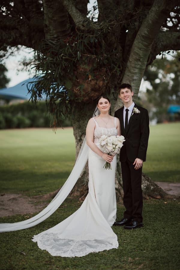 The bride Terion in a white wedding gown holding a bouquet of white flowers stands next to the groom Tyler in a black suit and tie, posing together in front of a large tree at Sandstone Point Hotel.