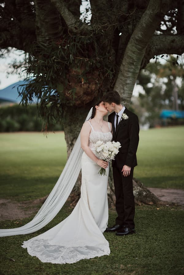 The bride Terion and groom Tyler share a kiss while standing in front of a large tree at Sandstone Point Hotelallo. Terion wears a white wedding gown with a long veil and holds a bouquet of white flowers. Tyler is dressed in a black suit with a white boutonniere.