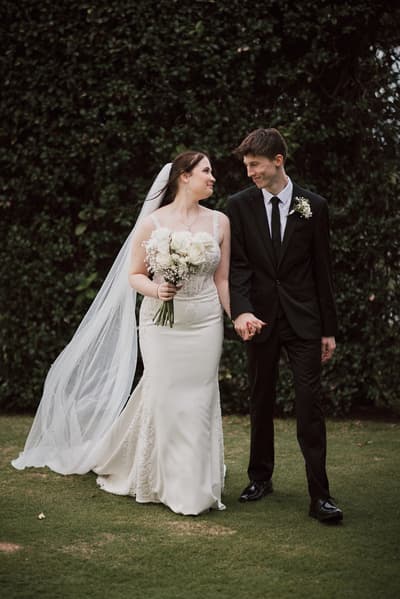Terion the bride in a white wedding gown holding a bouquet of white flowers and Tyler the groom in a black suit holding hands, standing on grass with a leafy hedge background. Brisbane elopement photographer by She Said Yes.