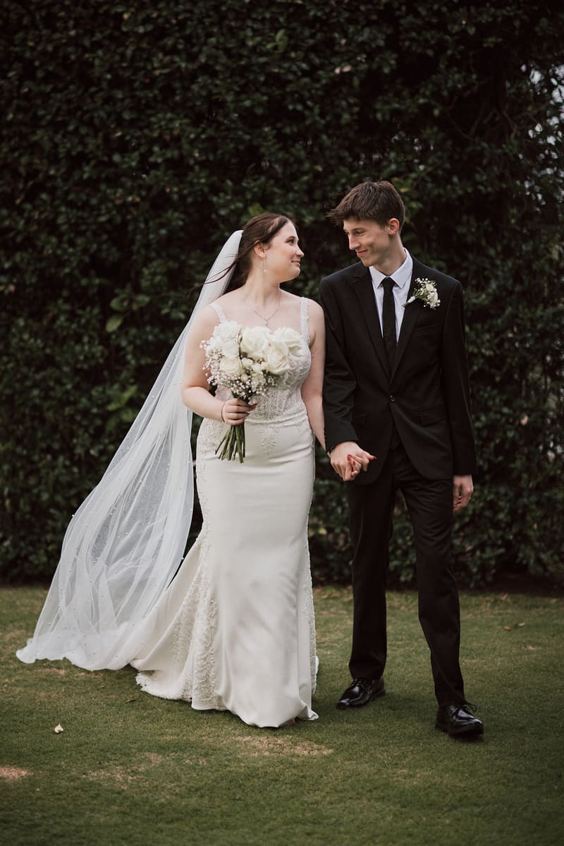 Terion the bride in a white wedding gown holding a bouquet of white flowers and Tyler the groom in a black suit holding hands, standing on grass with a leafy hedge background. Lead follow-up communication delay by She Said Yes.