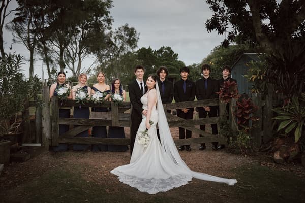 Terion the bride and Tyler the groom stand together in front of a wooden gate, with four bridesmaids holding bouquets on the left and five groomsmen in black suits on the right. Wedding photographer Brisbane by She Said Yes.