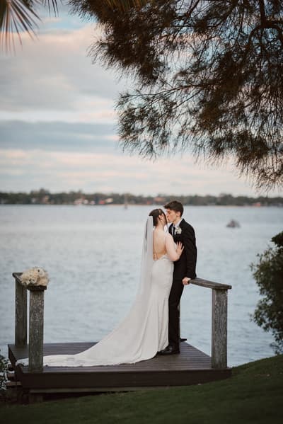 Terion the bride and Tyler the groom share a kiss on a wooden platform by the water. Sandstone point hotel wedding by She Said Yes.