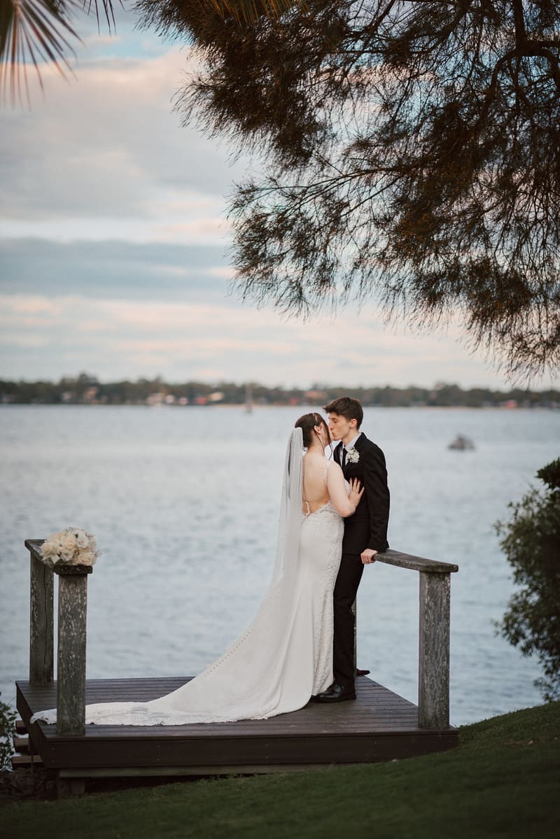 Terion the bride and Tyler the groom share a kiss on a wooden platform by the water. Sandstone point hotel wedding by She Said Yes.
