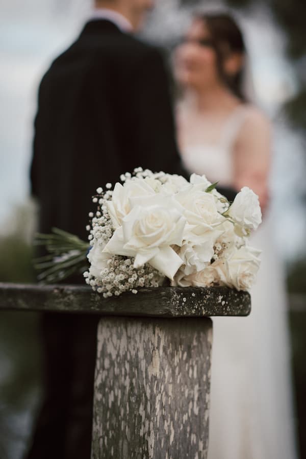 A bridal bouquet of white roses and baby's breath rests on a weathered wooden railing with the bride and groom blurred in the background at Sandstone Point Hotelallo.