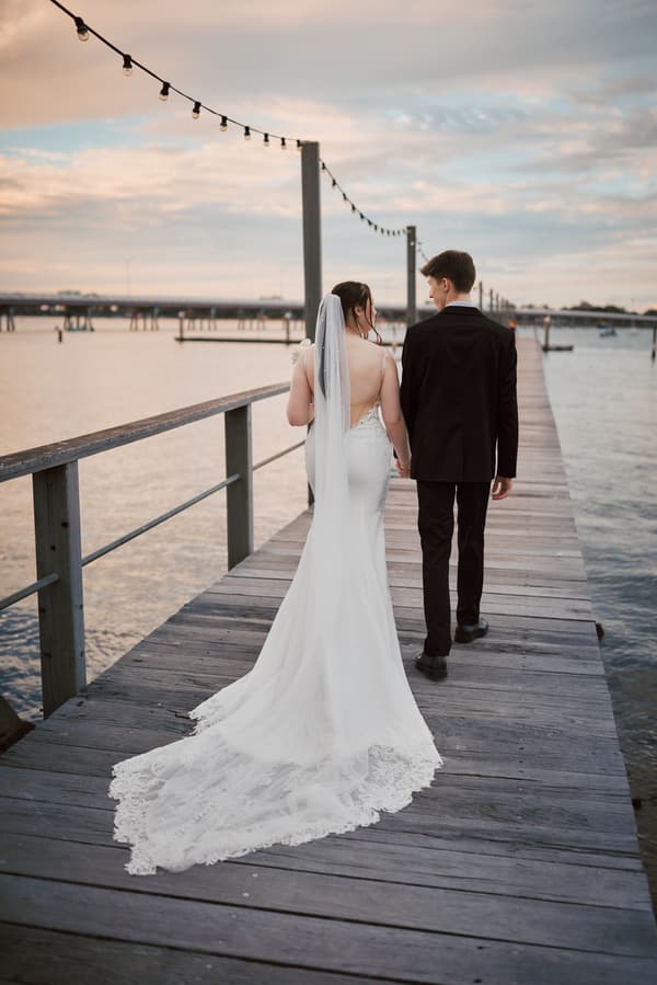 The bride Terion in a white wedding gown with a long lace train and veil, and the groom Tyler in a black suit, walk hand in hand along a wooden pier at Sandstone Point Hotel with water and a bridge in the background during sunset.