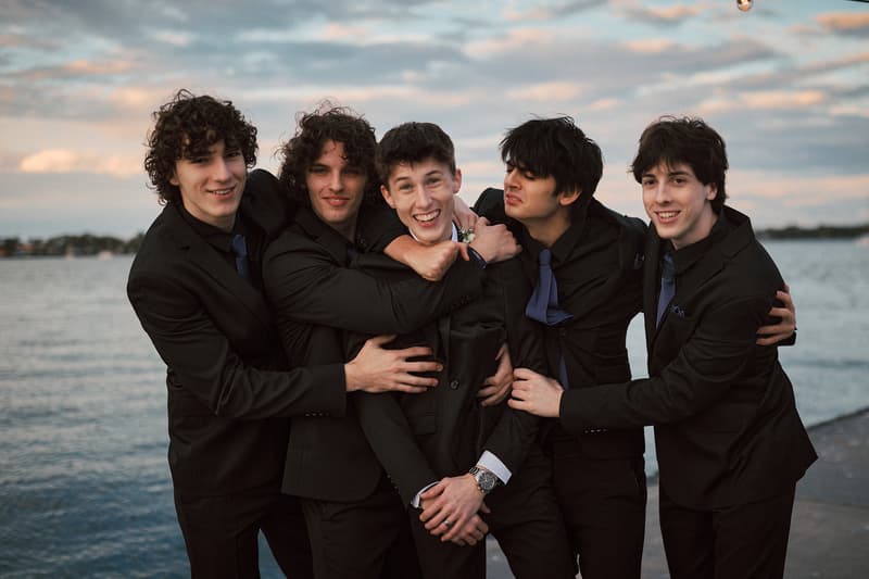 Five young men in black suits with blue ties pose together near the water at Sandstone Point Hotelallo during the couple portraits stage.